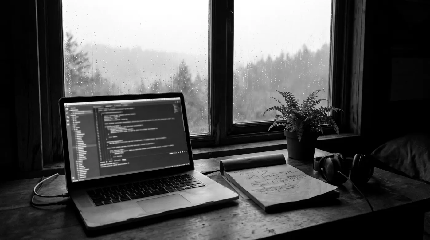 A desk by a rain-streaked window at dawn with an open laptop showing code, a notebook of sketched diagrams, headphones and a small plant.