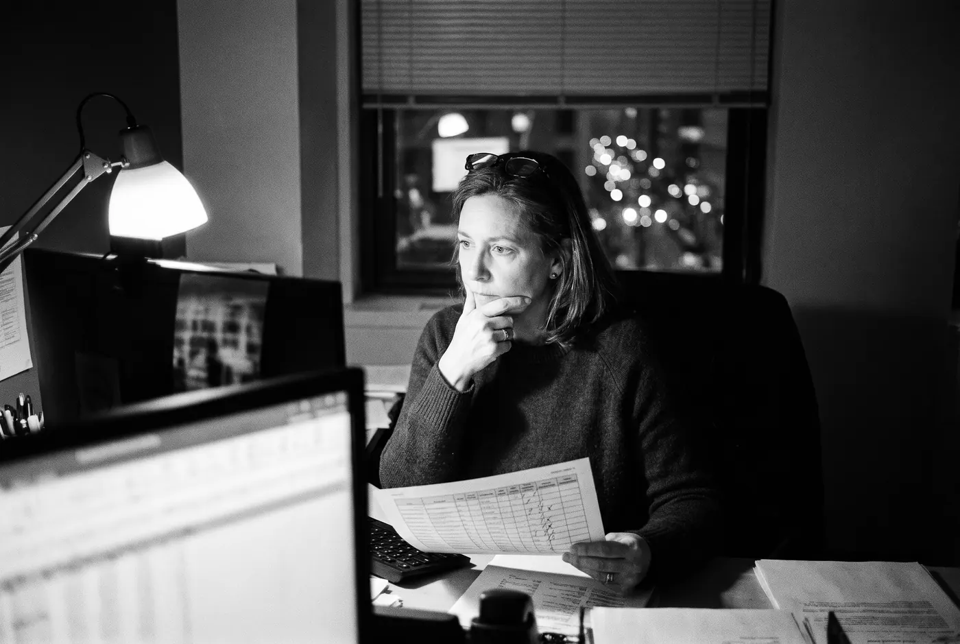 An analyst sits at her desk in the evening under a single lamp, holding a printed spreadsheet marked with highlighter.