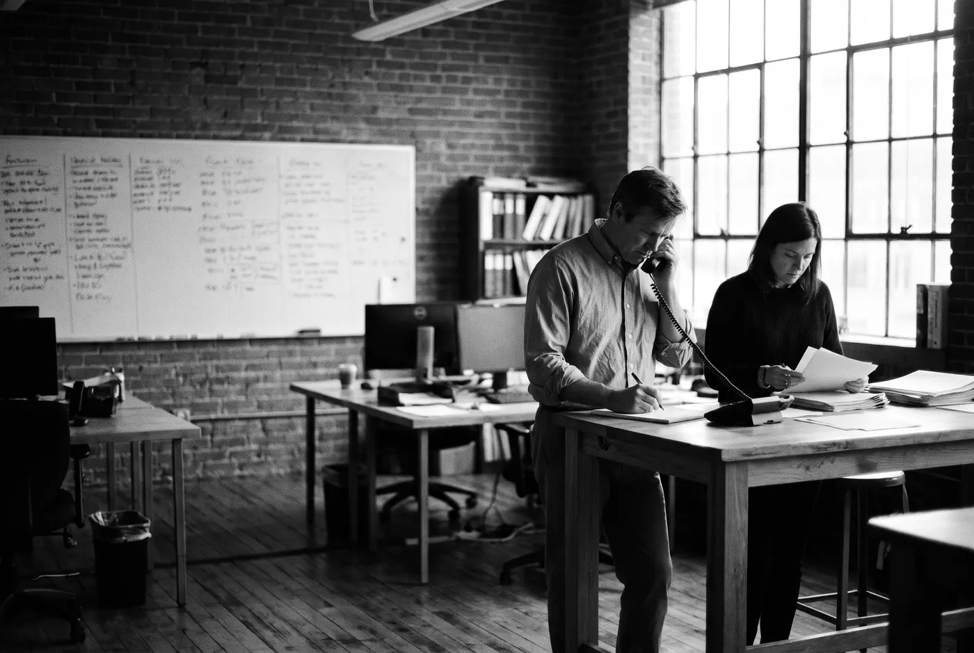 In a brick loft office, a man on a desk phone writes notes on a pad while a colleague beside him reads a printout.