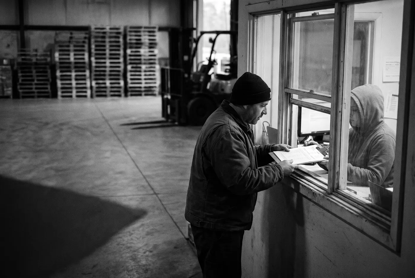 At a warehouse dispatch window, a man in a work jacket hands paperwork through to a colleague in the office, with pallets and a forklift behind him.